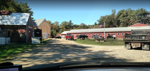 A dirt road leads through a farm with red barns, parked trucks, and an American flag in the background.