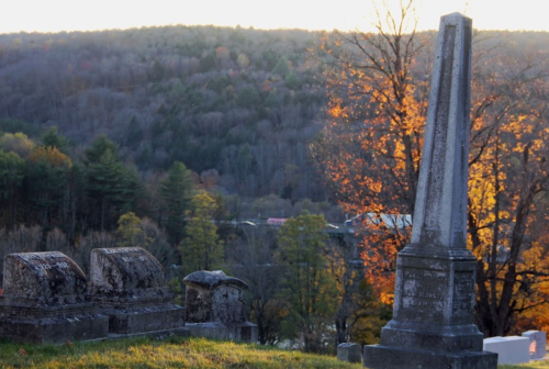 A cemetery scene featuring a tall obelisk gravestone and smaller headstones, with autumn trees and hills in the background.
