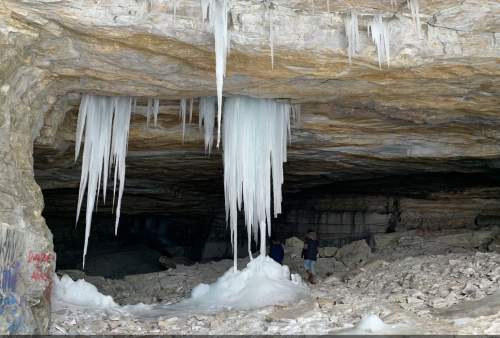 Icicles hang from the ceiling of a cave, with a rocky floor and two people standing in the background.