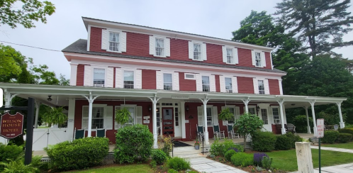 A red and white three-story hotel with a porch, surrounded by greenery and trees on a cloudy day.