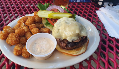 A plate with a cheeseburger topped with onions, lettuce, tomato, pickles, and a side of tater tots and dipping sauce.