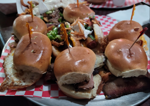 A platter of mini burgers with various toppings, garnished with toothpicks, served on a checkered paper.