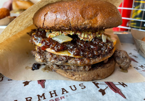 Close-up of a hearty burger with layers of beef, mustard, and a rich sauce, served on a brown bun.