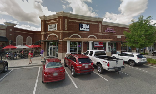 A brick building with "Groove" signage, outdoor seating, and parked cars in front on a sunny day.