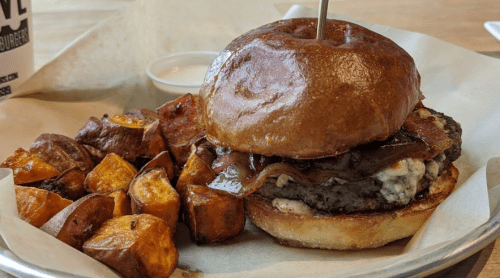 A juicy burger with bacon and blue cheese on a bun, served with crispy roasted sweet potato cubes and dipping sauce.