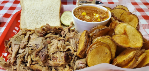 A plate featuring pulled pork, a slice of bread, potato chips, and a cup of chili with corn, all on a checkered tablecloth.