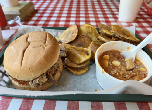 A plate with a pulled pork sandwich, crispy potato chips, and a bowl of chili on a red and white checkered tablecloth.