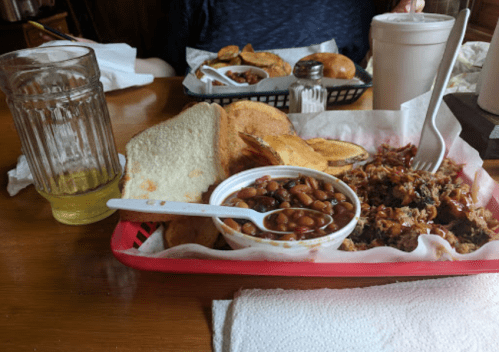 A red basket with pulled pork, baked beans, bread, and sides, accompanied by a glass and a drink in a cup.