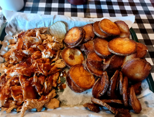 A plate of shredded barbecue chicken with pickles and a side of crispy potato wedges on a checkered tablecloth.