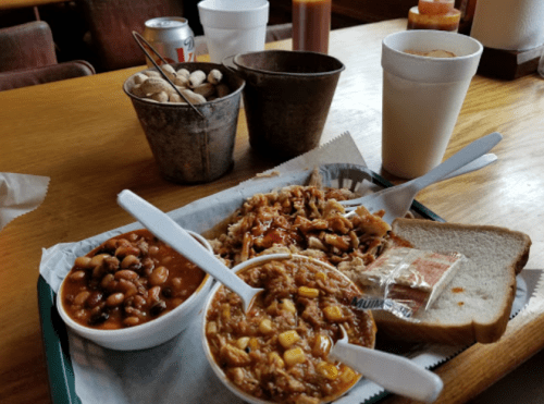 A plate with pulled pork, baked beans, corn dish, and bread, accompanied by drinks and peanuts in metal buckets.