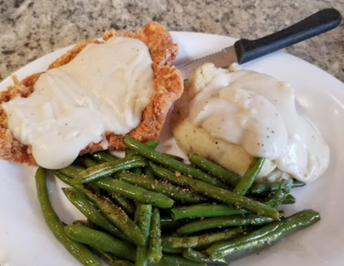 A plate with fried chicken topped with gravy, mashed potatoes with gravy, and seasoned green beans.