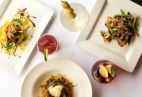 A top-down view of three gourmet dishes and colorful cocktails on a white tablecloth.