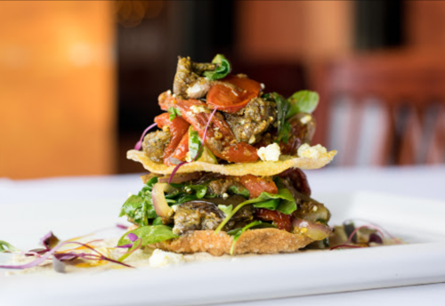 A colorful layered dish with greens, tomatoes, and crispy elements, elegantly presented on a white plate.