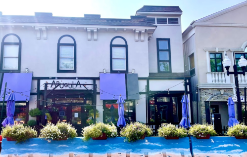A charming storefront with purple awnings, surrounded by greenery and outdoor seating, under a clear blue sky.