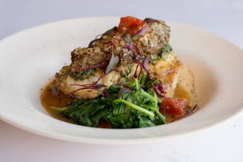A plate of baked fish topped with herbs, served with sautéed spinach and tomatoes in a light sauce.