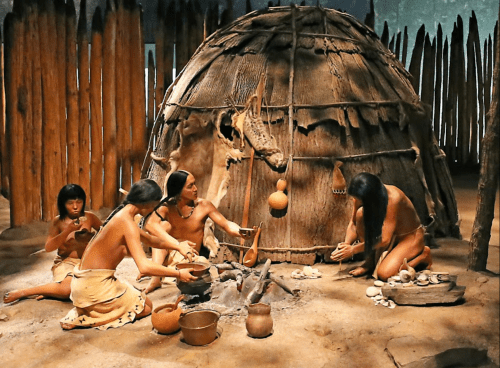 A group of Indigenous people cooking and crafting around a fire in front of a traditional dwelling.