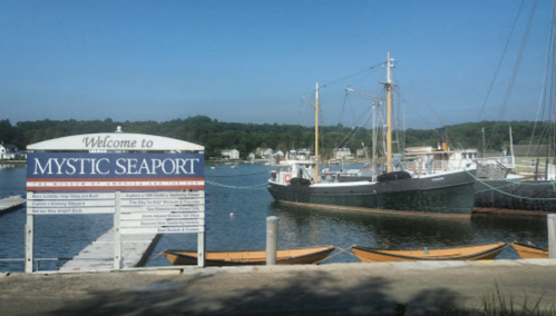 A welcome sign for Mystic Seaport with a docked boat and calm water in the background on a sunny day.