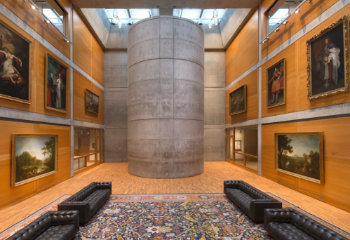 Interior of an art gallery featuring a central concrete column, wooden walls, and paintings displayed on the walls.