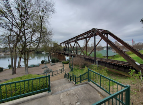 A rusty metal bridge spans a river, surrounded by trees and greenery, with steps leading down to the water.