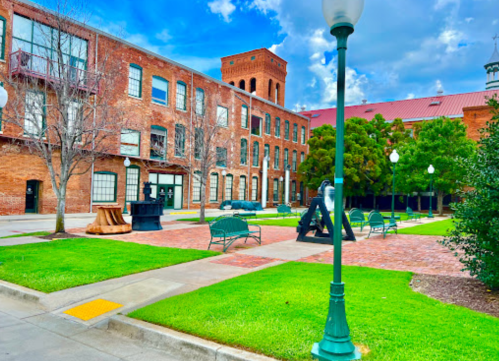 A brick courtyard with benches, sculptures, and green grass under a blue sky with clouds.