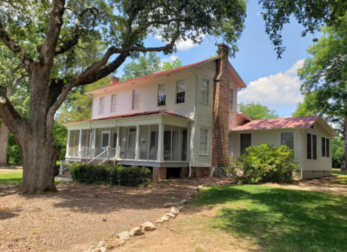 A two-story house with a red roof, surrounded by trees and greenery, featuring a porch and a well-maintained yard.
