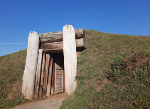 Ancient burial mound entrance with wooden beams, surrounded by green grass under a clear blue sky.