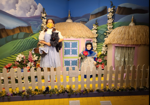 A diorama featuring a girl in a blue dress holding a dog, next to a doll, with thatched cottages and flowers in the background.