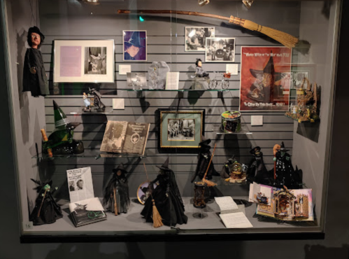 Display case featuring various witch-themed memorabilia, including dolls, books, and photographs.