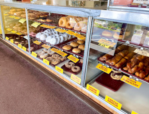 A display case filled with various donuts and pastries, labeled with prices and names.