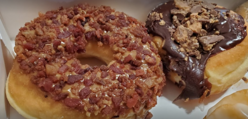 Two donuts in a box: one topped with chopped bacon and the other with chocolate and crushed candy.