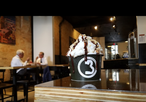 A close-up of a dessert cup with whipped cream and chocolate drizzle, with two people chatting in the background.
