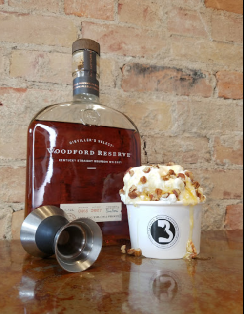 A bottle of Woodford Reserve bourbon next to a cup of ice cream topped with caramel and popcorn, with jigger tools nearby.