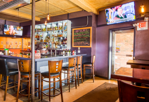 A cozy bar interior with wooden stools, shelves of liquor, and TVs displaying sports, illuminated by warm lighting.
