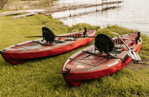 Two red kayaks are parked on green grass by a calm lake, with a wooden dock visible in the background.