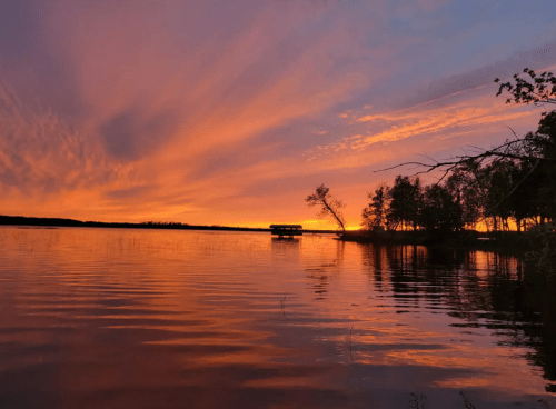 A serene lake at sunset, with vibrant orange and purple skies reflecting on the water and silhouettes of trees.