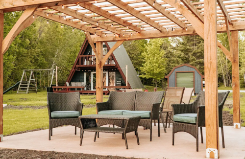 A cozy outdoor seating area with wicker furniture under a wooden pergola, overlooking a green yard and A-frame cabin.