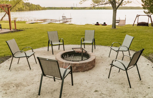 A circular fire pit surrounded by six chairs, overlooking a calm lake and green grass.