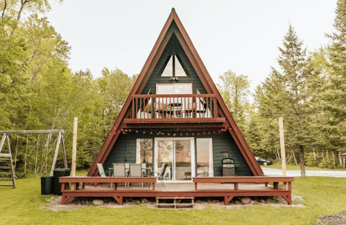 A-frame cabin with a wooden deck, surrounded by trees and a grassy area, featuring a balcony and large windows.
