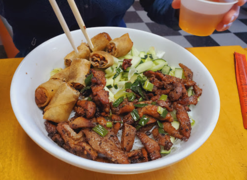 A bowl of stir-fried meat with green onions, served with spring rolls and cucumber slices, alongside a drink.