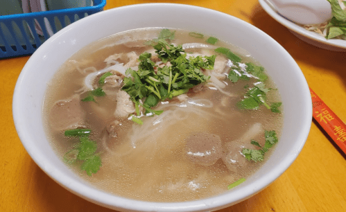A bowl of soup with noodles, meat, and fresh herbs, served on a wooden table.