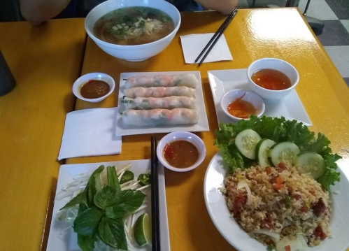A table set with a bowl of soup, spring rolls, fried rice, fresh herbs, and dipping sauces on a bright yellow surface.