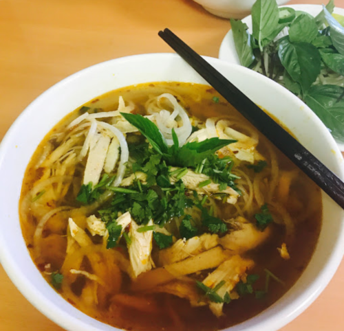 A bowl of noodle soup topped with shredded chicken, herbs, and green onions, with chopsticks resting on the side.