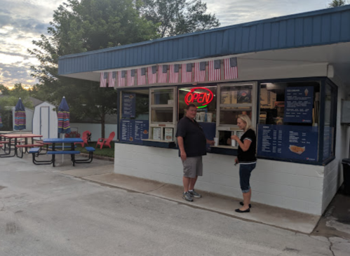 A man and woman stand at an outdoor food stand with an "OPEN" sign, surrounded by picnic tables and flags.