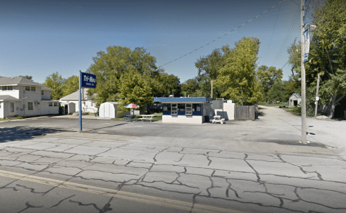 A small roadside building with a blue roof and sign, surrounded by trees and a cracked pavement road.