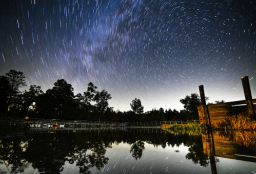 Star trails swirl above a calm lake, reflecting trees and a wooden structure in the foreground.