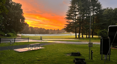 A vibrant sunset over a grassy field, framed by trees, with a picnic area and a hint of mist in the air.
