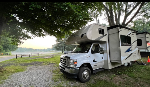 A parked RV on a gravel site surrounded by trees, with a misty landscape in the background.