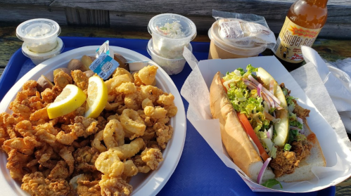A plate of fried seafood with lemon slices, alongside a seafood sandwich topped with fresh vegetables and a drink.