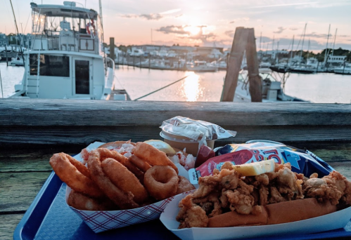 A scenic view of a marina at sunset with a tray of fried seafood and onion rings in the foreground.