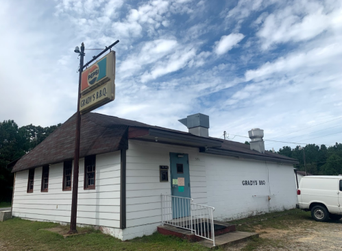 A small, white BBQ restaurant with a sign reading "Grady's BBQ" and a blue door, set against a cloudy sky.